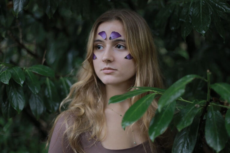 Portrait of a woman with long blonde hair and purple flower petals on her cheeks, standing in front of a tree with green leaves.